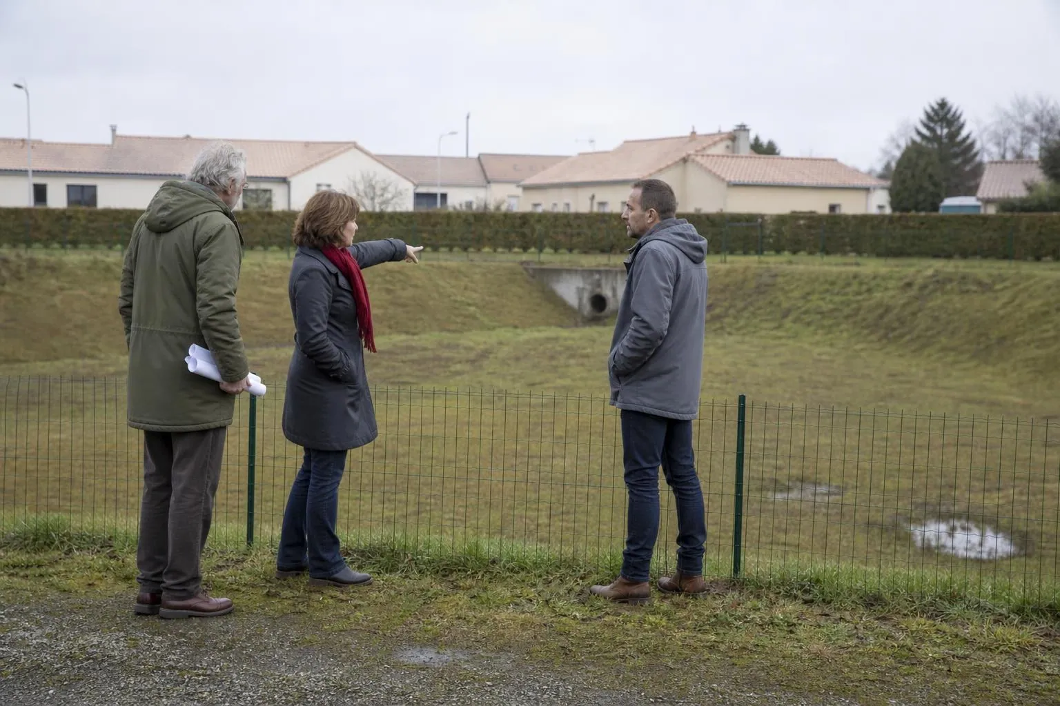 Trois personnes discutent debout au bord d’un bassin de rétention enherbé, l’une tenant un plan roulé sous le bras